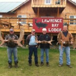 Four men stand in front of Lawrence Bay Lodge holding large antlers.