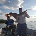 Two people on a boat; one holds a large fish, and the other takes a photo. The sky is partly cloudy.