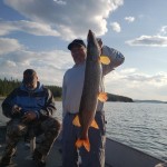 Two people on a boat; one holds a large fish while the other looks down at a device. Lake and sky in the background.