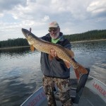 Man holding a large fish on a boat with a lake and forest in the background.