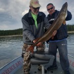 Two men on a boat holding a large fish they caught, with a lake and forested shoreline in the background.