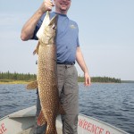 Man on a boat holding a large fish vertically by the mouth in one hand, with water and trees in the background.