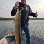 Person on a boat holding a large fish with one hand, giving a thumbs up with the other, under a partly cloudy sky.