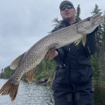 Person holding a large fish on a boat, with trees and a cloudy sky in the background.