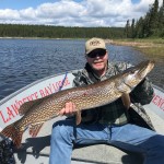 Person seated in a boat holding a large fish, with a lake and forest in the background under a partly cloudy sky.