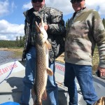 Two men on a boat holding a large fish, with trees and water in the background under a partly cloudy sky.