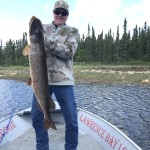 Person in camo sweater and cap holding a large fish on a boat, with trees and water in the background.