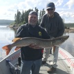 Two people on a boat, one holding a large fish. Trees and a cloudy sky are in the background.