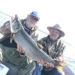 Two men on a boat holding a large fish. The man on the left wears a blue cap; the man on the right wears a wide-brimmed hat.