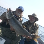Two men sitting in a boat, holding a large fish they caught on a lake.