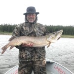 Person in camouflage clothing holding a large fish on a boat, with a forested shoreline in the background.