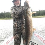 Person in camo outfit holding a large fish on a boat with a forest and water in the background.