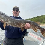 Person holding a large fish on a boat in a lake, with forested shoreline in the background.