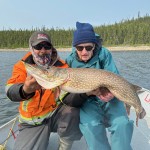 Two people in a boat holding a large fish, with a forested shoreline in the background.