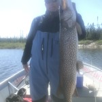 Person in a blue outfit holding a large fish on a boat with trees and water in the background.