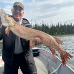 Person smiling while holding a large fish on a boat, with trees and water in the background.
