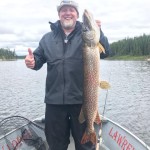 Person on a boat smiling, holding a large fish in one hand and giving a thumbs-up with the other, near a wooded riverbank.