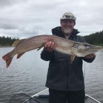 Man standing on a boat holds a large fish with a lake and forest in the background under a cloudy sky.