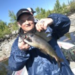 Person in a blue jacket holding a fish on a boat, with rocks and trees in the background under a clear sky.