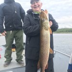 Two people on a boat, one holding a large fish and smiling. The other stands nearby in a hat and waterproof gear.