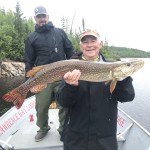 Two people on a boat; one holds a large fish, possibly a pike. Trees and water are in the background.