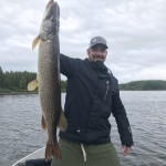 Person in a boat holds a large fish vertically. Overcast sky and tree-lined shoreline in the background.