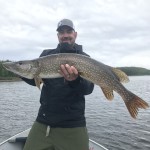 Person holding a large fish on a boat in a lake, wearing a black jacket and cap, with a forested shoreline in the background.
