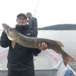 Man on a boat holding a large fish, with another person in the background. Overcast sky and calm water surround them.