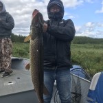 Person holding a large fish on a boat, wearing a hooded jacket and jeans, with another person in the background.