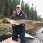Person standing in a boat holding a large fish, with a forested shoreline in the background.