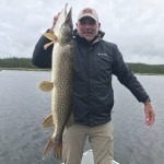 Person standing on a boat holding a large fish vertically, wearing a dark jacket and cap, with a cloudy sky and trees in the background.
