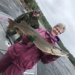 Woman in a pink raincoat holding a large fish on a boat, with a man in camouflage behind her, overcast sky in the background.