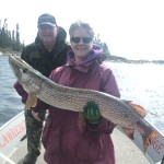 Two people on a boat holding a large fish. The woman in front is smiling, wearing a purple jacket and gloves.