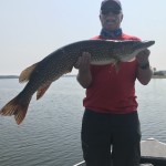 Person in red shirt and cap holding a large fish on a boat, with a calm body of water in the background.