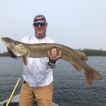 Man holding a large fish on a boat, with water and trees in the background.