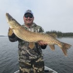 Man in camo outfit holding a large fish on a boat with a lake and trees in the background.