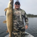 Person in camouflage clothing on a boat holding a large fish upright, with a forested shoreline in the background.