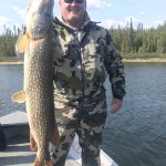 Smiling man in camouflage outfit holding a large fish on a boat, with forested shoreline in the background.