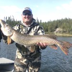 Man in camouflage holds large fish on a boat with a lake and forest in the background under a clear sky.