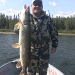 Man in camouflage clothing holds a large fish on a boat, with trees and water in the background.