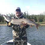 Smiling person in camouflage gear holds a large fish on a boat, with a forested shoreline in the background under a clear sky.