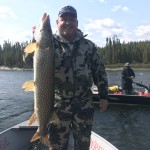 Person on a boat holding a large fish, another person visible on a separate boat in the background, surrounded by water and trees.