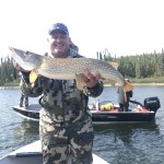 Man in camouflage holds a large fish on a boat, with another boat and pine trees in the background.