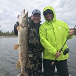 Two people on a boat, one holding a large fish and the other holding a fishing rod, smiling at the camera.