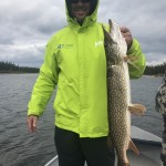 Person in a bright green jacket holding a large fish on a boat in a lake with overcast skies.