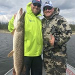 Two people on a boat, one holding a large fish, both smiling and wearing outdoor jackets and caps, with a cloudy sky in the background.