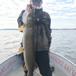 Person standing on a boat holding a large fish vertically with two hands, wearing a jacket, hat, and sunglasses.