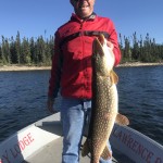 Person in a red jacket and cap stands on a boat holding a large fish, with a lake and trees in the background.