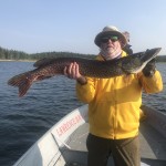 Person in yellow jacket holding a large fish on a boat, with a lake and trees in the background.