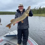 Man on a boat holding a large fish, smiling. Overcast sky and trees in the background. Fishing net on the boat's floor.